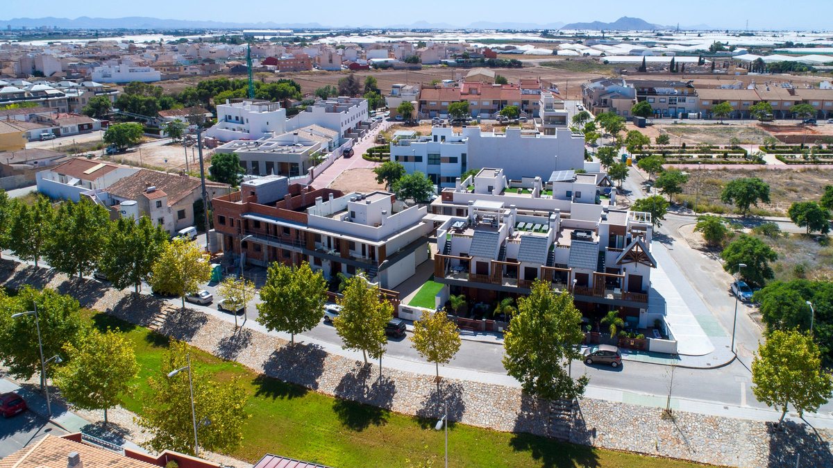 Aerial view of the quiet residential area in Pilar de la Horadada: apartments with roof terraces, trees along a green strip and the farmland and hills beyond